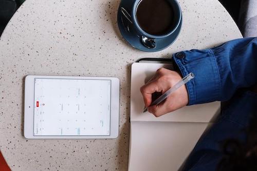 A person writing on a journal, looking at a calendar on an iPad